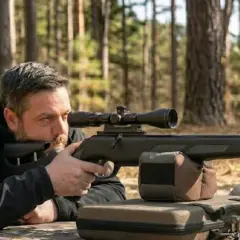 Un homme en tenue noire, couché dans une forêt, vise avec une carabine équipée d'une lunette. Le kit de puissance 40 joules est prêt pour le tir.