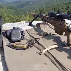 Carabine de précision beige, lunette, télémètre et sac de transport sur une table en bois, avec une forêt en arrière-plan.