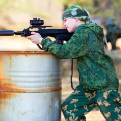 Un jeune garçon en tenue de camouflage vise avec un fusil laser, caché derrière un baril rouillé. Une partie de jeu de tir sur terrain privé.