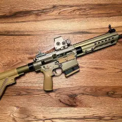 Un fusil HK MR308 couleur sable, avec lunette, repose sur un fond en bois.