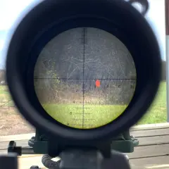 Lunette de tir visant une cible rouge. La scène extérieure montre un champ et des arbres. On peut imaginer la vitesse balle 9mm.