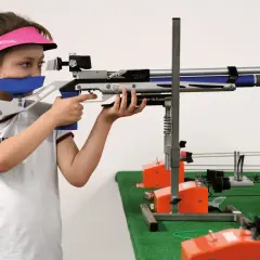 Jeune fille concentrée, vise avec une carabine de tir. Elle porte une visière rose et des protections auditives. Elle est prête pour sa licence de tir.