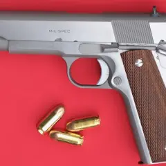 Un pistolet MIL-SPEC argenté avec des plaquettes de crosse en bois et trois cartouches à blanc à côté.
