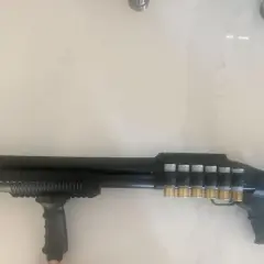 Un fusil Maverick 88 tactique, noir, avec une poignée avant, un rail supérieur et des cartouches dorées visibles sur le côté.