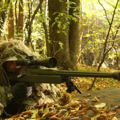Un tireur d'élite camouflé dans les bois, tenant un fusil Macmillan.
