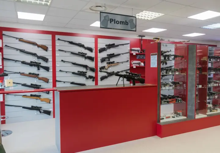Vitrines d'un magasin d'armes présentant des fusils et carabines, dont un pistolet à bille interdit, sur des murs rouges et gris.