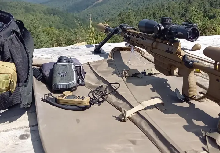 Carabine de précision beige, lunette, télémètre et sac de transport sur une table en bois, avec une forêt en arrière-plan.