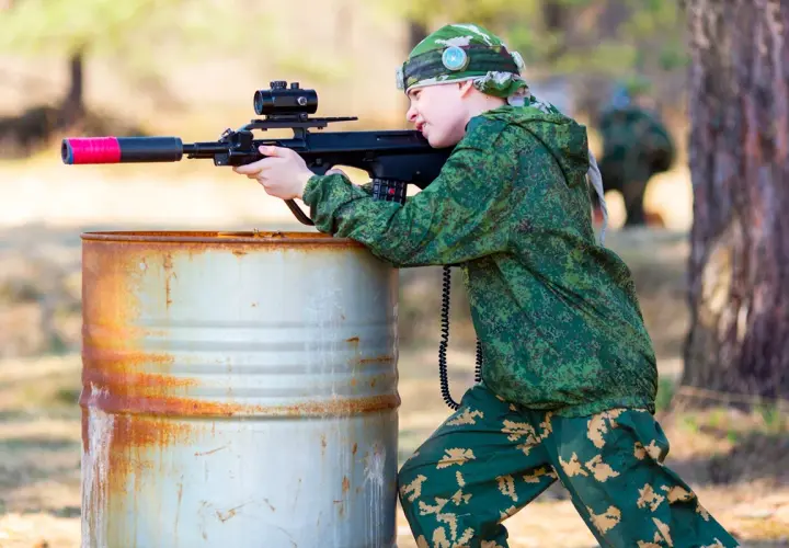 Un jeune garçon en tenue de camouflage vise avec un fusil laser, caché derrière un baril rouillé. Une partie de jeu de tir sur terrain privé.