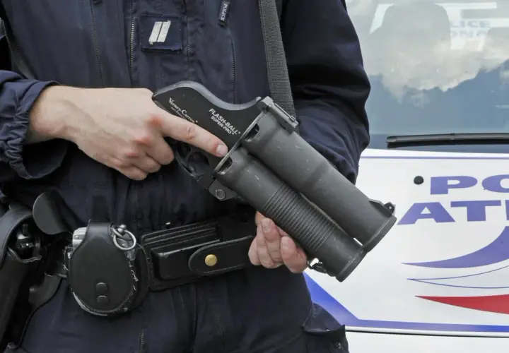 Un policier tient un lanceur flashball, arme non létale, près d'une voiture de police.