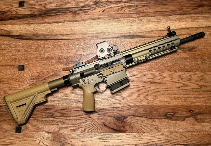 Un fusil HK MR308 couleur sable, avec lunette, repose sur un fond en bois.