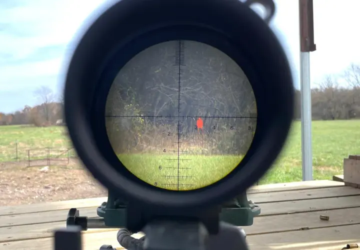 Lunette de tir visant une cible rouge. La scène extérieure montre un champ et des arbres. On peut imaginer la vitesse balle 9mm.