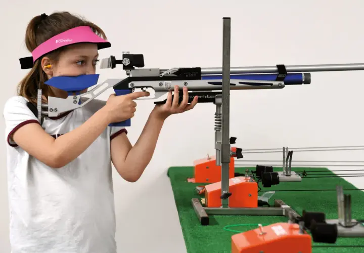 Jeune fille concentrée, vise avec une carabine de tir. Elle porte une visière rose et des protections auditives. Elle est prête pour sa licence de tir.