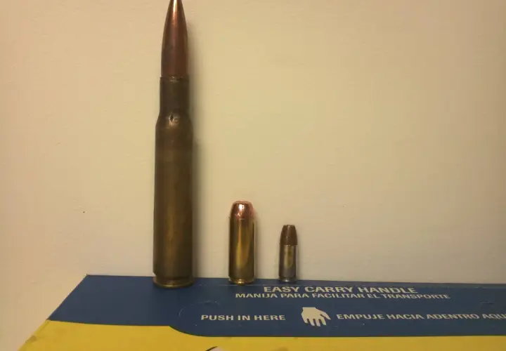 Trois cartouches de tailles différentes, dont une imposante de calibre 50 en mm, posées sur une boîte bleue et jaune.