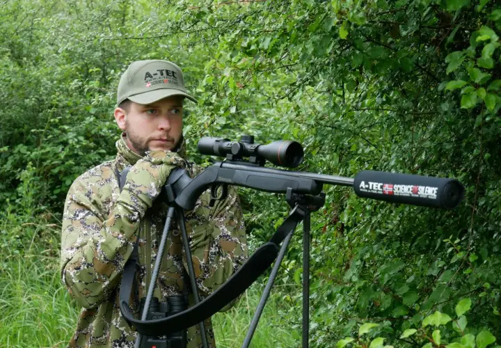Chasseur en camouflage, prêt à l'action avec sa carabine A-Tec, la meilleure carabine pour battue sanglier, dans un sous-bois dense.