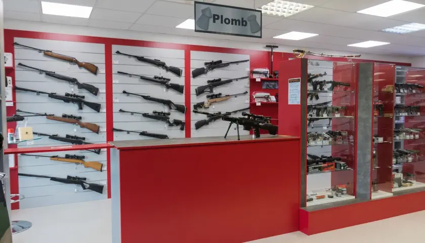Vitrines d'un magasin d'armes présentant des fusils et carabines, dont un pistolet à bille interdit, sur des murs rouges et gris.