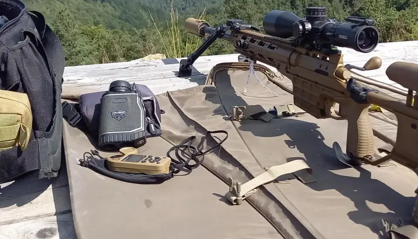 Carabine de précision beige, lunette, télémètre et sac de transport sur une table en bois, avec une forêt en arrière-plan.