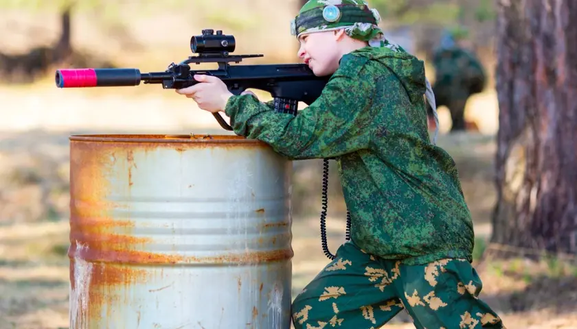 Un jeune garçon en tenue de camouflage vise avec un fusil laser, caché derrière un baril rouillé. Une partie de jeu de tir sur terrain privé.