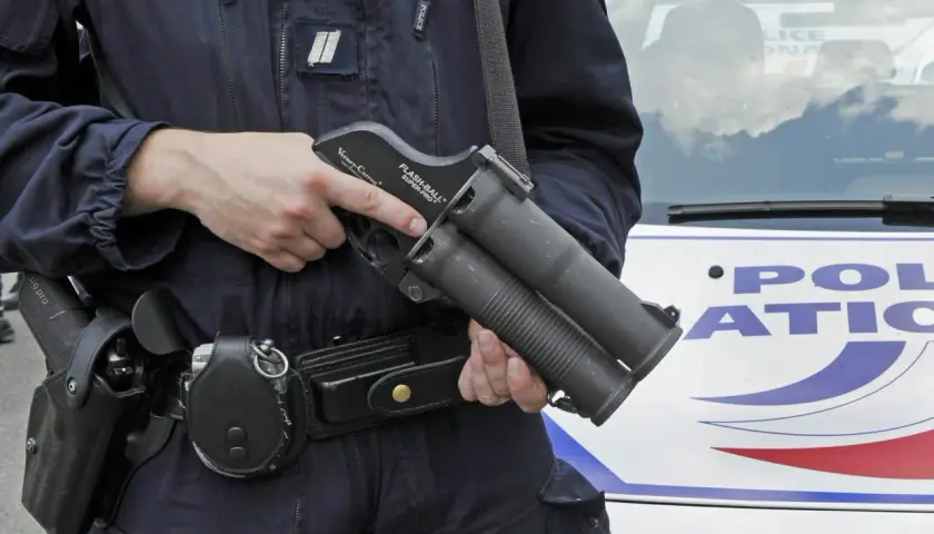 Un policier tient un lanceur flashball, arme non létale, près d'une voiture de police.