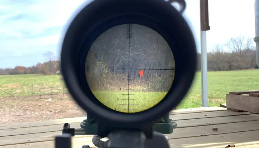 Lunette de tir visant une cible rouge. La scène extérieure montre un champ et des arbres. On peut imaginer la vitesse balle 9mm.
