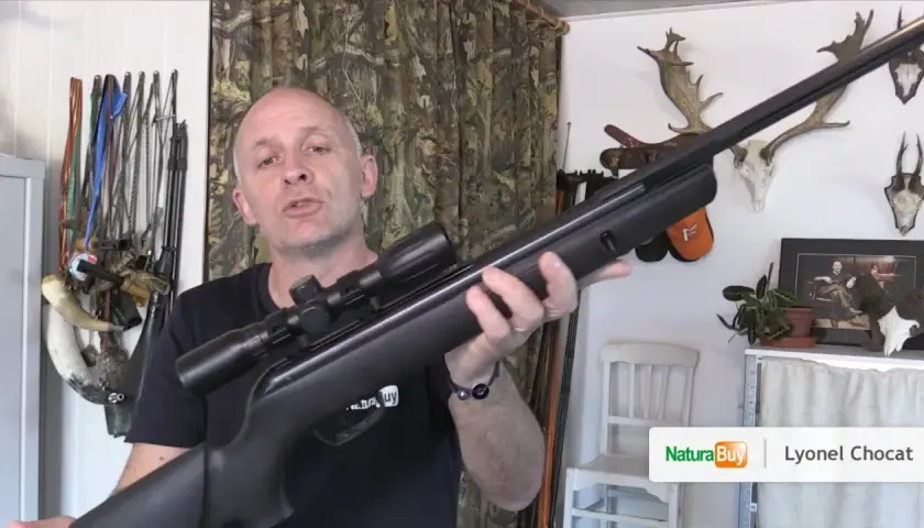 Un homme présente une carabine à air comprimé de 20 joules, avec des trophées de chasse en arrière-plan.