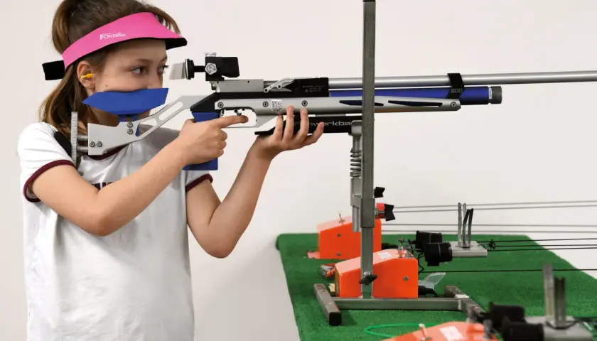 Jeune fille concentrée, vise avec une carabine de tir. Elle porte une visière rose et des protections auditives. Elle est prête pour sa licence de tir.
