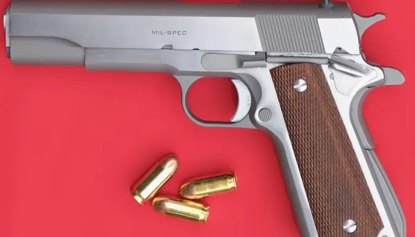 Un pistolet MIL-SPEC argenté avec des plaquettes de crosse en bois et trois cartouches à blanc à côté.