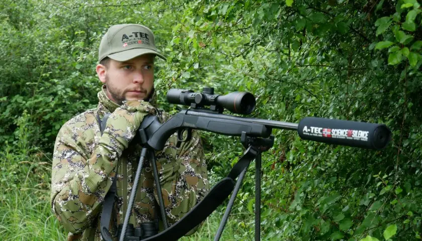 Chasseur en camouflage, prêt à l'action avec sa carabine A-Tec, la meilleure carabine pour battue sanglier, dans un sous-bois dense.