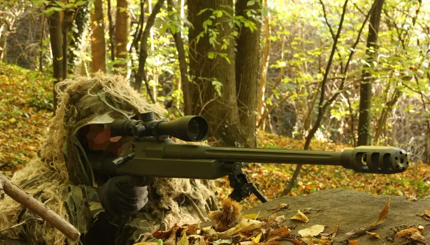 Un tireur d'élite camouflé dans les bois, tenant un fusil Macmillan.