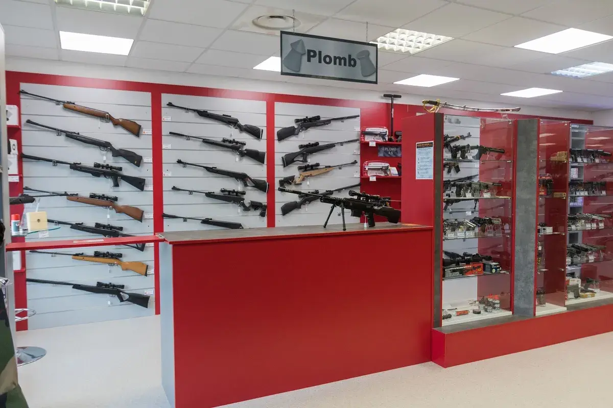 Vitrines d'un magasin d'armes présentant des fusils et carabines, dont un pistolet à bille interdit, sur des murs rouges et gris.