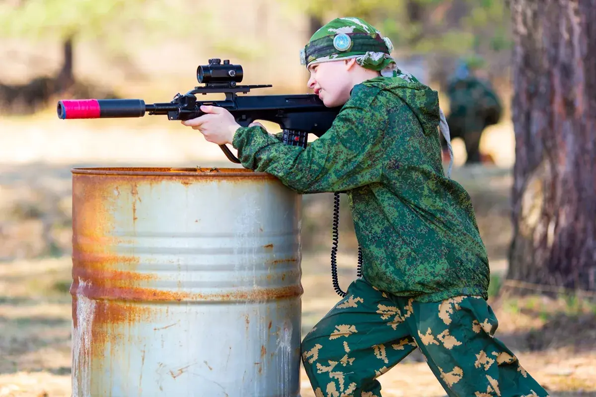 Un jeune garçon en tenue de camouflage vise avec un fusil laser, caché derrière un baril rouillé. Une partie de jeu de tir sur terrain privé.