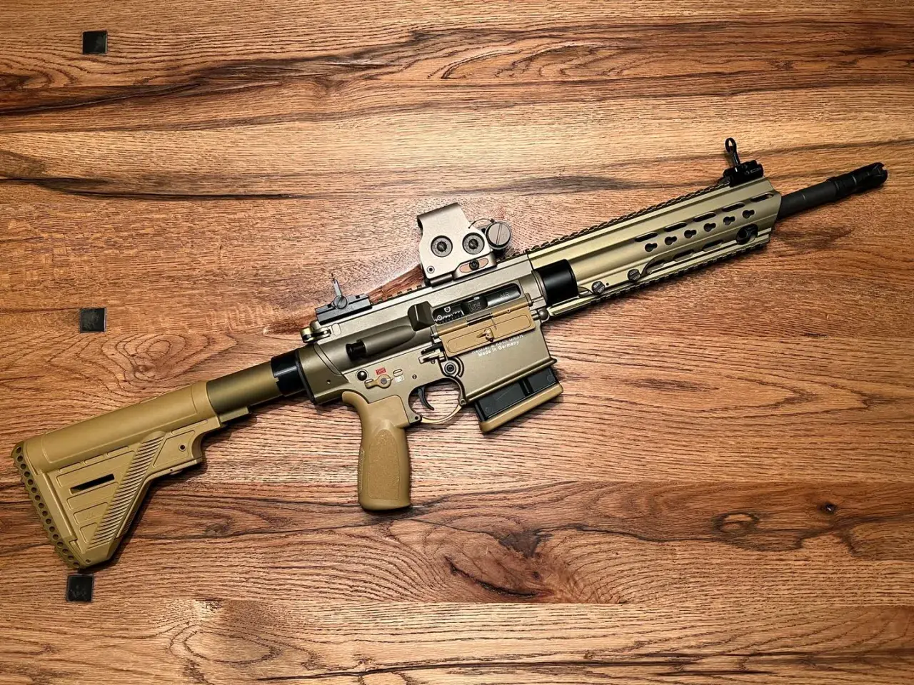 Un fusil HK MR308 couleur sable, avec lunette, repose sur un fond en bois.
