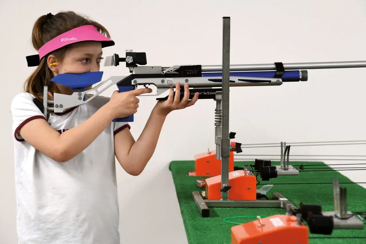 Jeune fille concentrée, vise avec une carabine de tir. Elle porte une visière rose et des protections auditives. Elle est prête pour sa licence de tir.