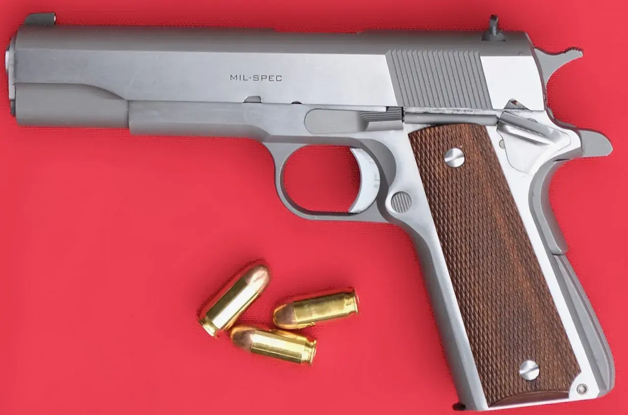 Un pistolet MIL-SPEC argenté avec des plaquettes de crosse en bois et trois cartouches à blanc à côté.