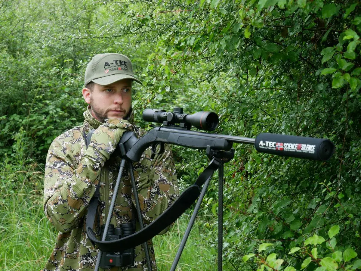 Chasseur en camouflage, prêt à l'action avec sa carabine A-Tec, la meilleure carabine pour battue sanglier, dans un sous-bois dense.