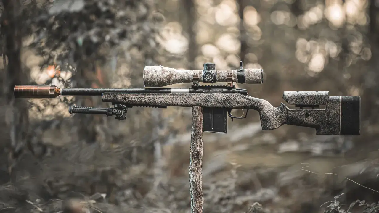 Un fusil Macmillan, prêt pour la chasse, avec lunette et silencieux, se dresse sur un support en bois dans un décor forestier.