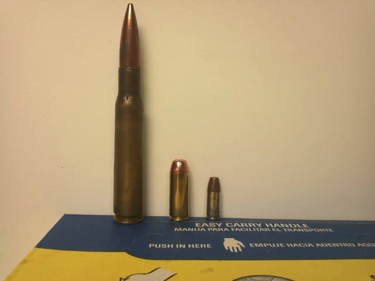 Trois cartouches de tailles différentes, dont une impressionnante calibre 50, posées sur une boîte bleue et jaune.
