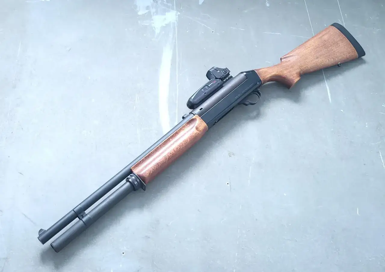 Fusil Benelli 121 avec viseur reflex, bois sombre et canon noir, posé sur une surface grise.